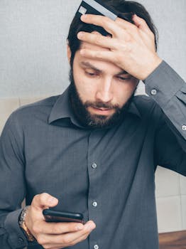 A stressed man looks at his smartphone, holding a credit card in his hand.