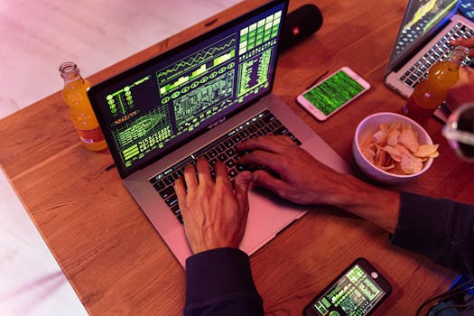 Hands typing on a laptop with coding, phone on desk, symbolizing cybersecurity.