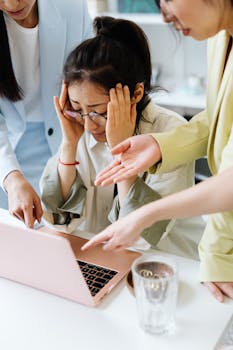 An adult woman looking stressed at work, surrounded by coworkers pointing at a laptop indoors.